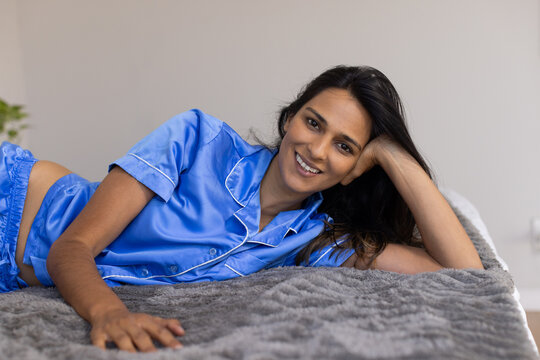 Woman reclining in bedroom, smiling and propping head, wearing blue satin pajamas on gray blanket