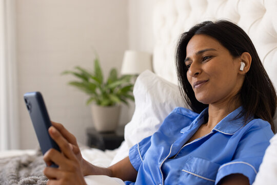Adult female reclining in bed wearing blue pajama top, holding smartphone and earbuds, copy space
