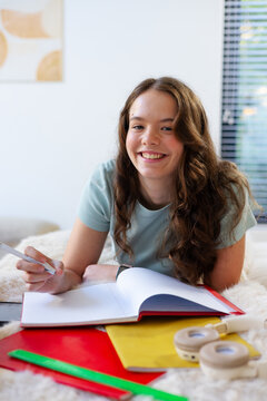 Teenage female lying on bed, smiling while writing in notebook with pen, folders and ruler nearby