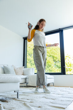 Woman dancing, holding smartphone, wearing pink headphones and raglan shirt in living room