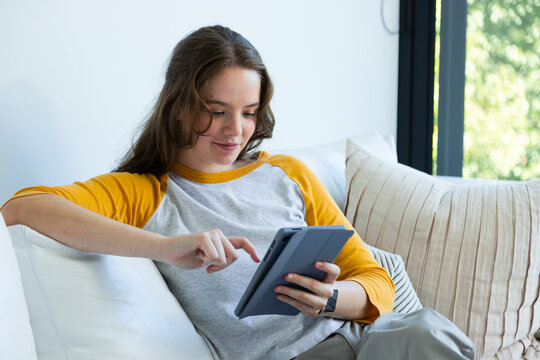 Adult female reclining on couch at home with pillows in raglan yellow shirt, tapping gray tablet
