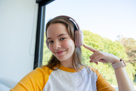 Female in twenties wearing yellow raglan shirt and pink headphones, pointing at earcup by window