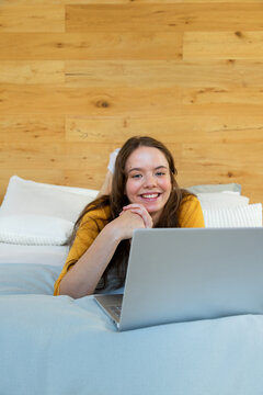 Female lying on stomach on bed wearing mustard yellow top, smiling and typing on silver laptop