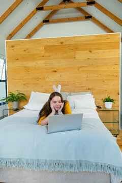 Adult female propping on elbows watching laptop in bedroom wearing mustard top and bunny headband