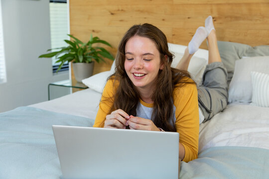 Woman lying on stomach on bed at home wearing yellow top and sweatpants, using silver laptop