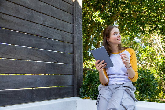 Female sitting on white concrete bench in garden corner, wearing yellow raglan, holding gray tablet