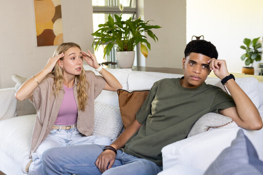 Diverse couple sitting on white sofa in living room with potted plant, both looking stressed