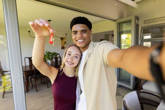 Diverse couple standing at home threshold, man taking selfie with phone, woman holding red key fob