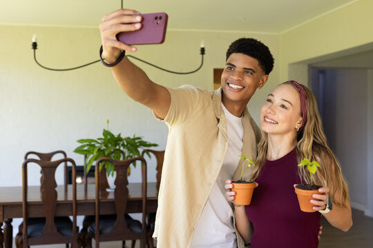 Couple taking selfie in dining area, man in beige shirt holding smartphone, woman holding clay pots