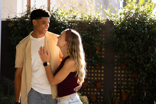 Diverse couple standing in sunlit backyard garden by wooden trellis, smiling, smartwatch visible
