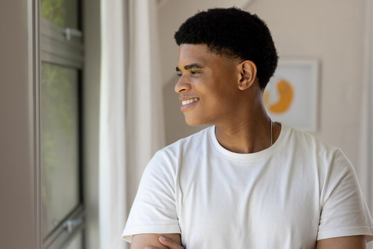 African American man gazing out window, wearing white tee silver chain, sheer curtains, copy space