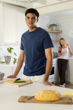 Diverse couple standing in bright kitchen, man in navy tee, woman using smartphone, loaf on board