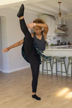 Adult African American woman wearing tank top and leggings doing high kick on kitchen wood floor