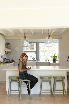 Woman sitting on sage-green bar stool at kitchen island wearing headphones and checking smartphone
