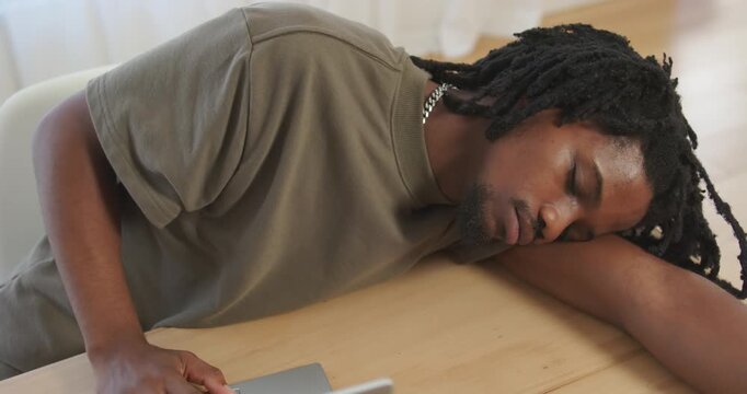Feeling tired, African American male resting head on forearm near silver device at wood table