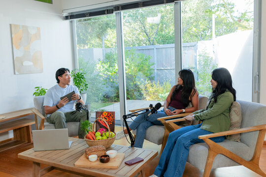 Diverse co-hosts sitting in living room, recording discussion with mic arms, tablet, smartphone