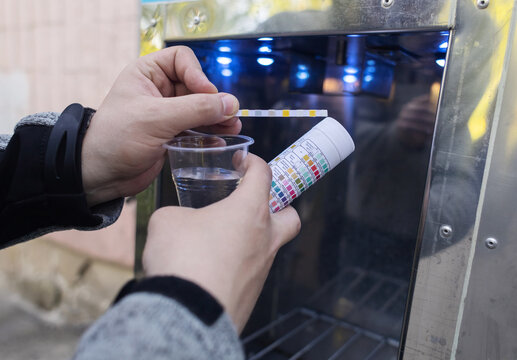 Man checking water quality at a public vending machine using a test strip. Analysis of purified drinking water in a plastic cup. Safety, pH, and hardness control of urban water supply.