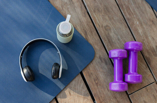 Blue mat, black headphones, water bottle, purple dumbbells are resting on wooden deck with shadows
