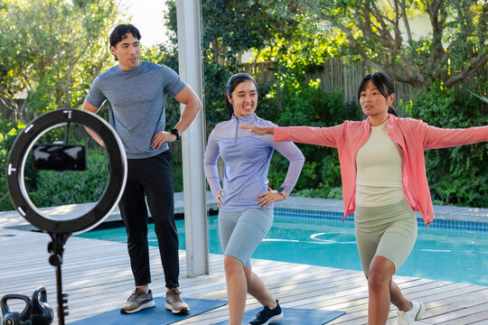 Diverse group in sportswear lunging on wooden deck beside pool with ring light phone and kettlebell