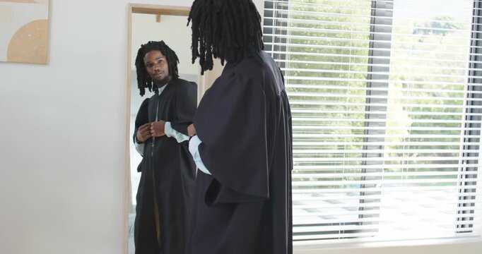 African American man fastening and smoothing ceremonial gown in bedroom mirror, preparing for event