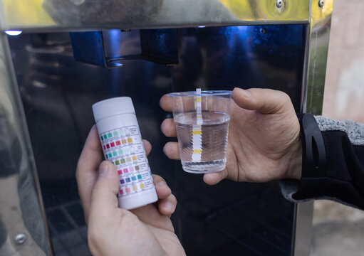 Man checking water quality at a public vending machine using a test strip. Analysis of purified drinking water in a plastic cup. Safety, pH, and hardness control of urban water supply.