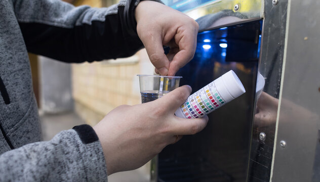 Man checking water quality at a public vending machine using a test strip. Analysis of purified drinking water in a plastic cup. Safety, pH, and hardness control of urban water supply.