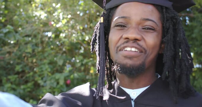 African male in cap and gown celebrating upon graduating smiling waving red ribbon scroll in garden