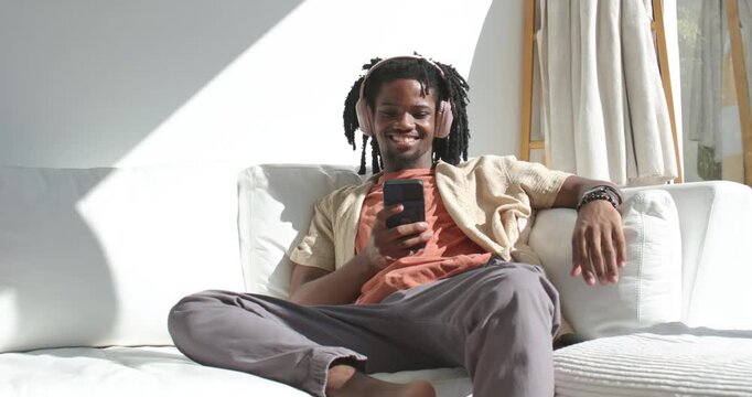 African American man sitting on white sofa with headphones holding phone and laughing at funny clip