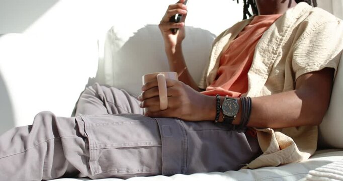 Adult African man settling back shifting phone viewing screen holding mug in rust tee on couch