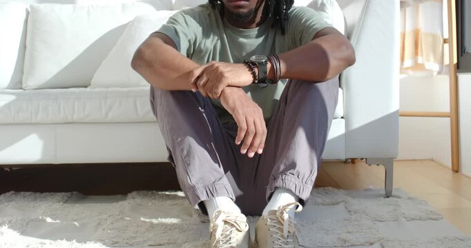 Adult African man sitting on rug by white sofa, adjusting wristwatch as sunlight crossing rug