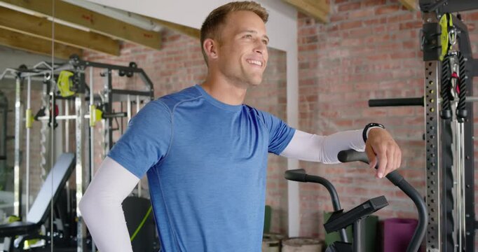 30s man leaning on elliptical in home gym, glancing up and smiling toward camera, wearing shirt