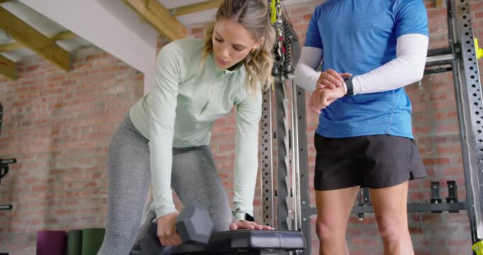 Trainer tapping smartwatch, cueing pace, client performing hex dumbbell rows on step bench in gym