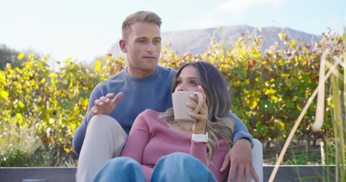 Diverse couple sitting on patio by vines, woman wearing watch leaning back sipping mug, he speaking