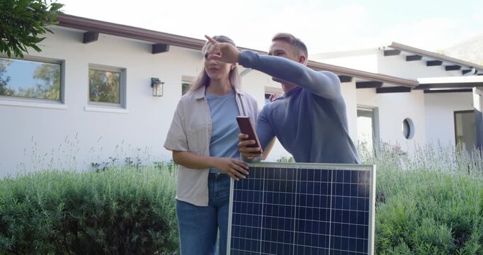 Diverse couple at modern house holding solar panel pointing to roof and checking phone for install