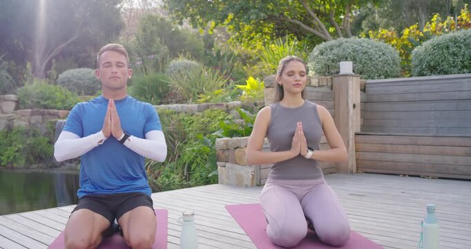 Couple in yoga wear kneeling palms together on deck mats beside pond, sunbeam appearing, meditating