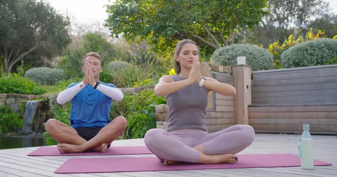 Pair wearing activewear doing yoga on mats on deck by waterfall, breathing, forming mudra for calm