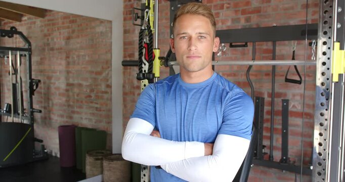 Adult male in blue top standing arms crossed, camera tightening revealing rack and straps in gym