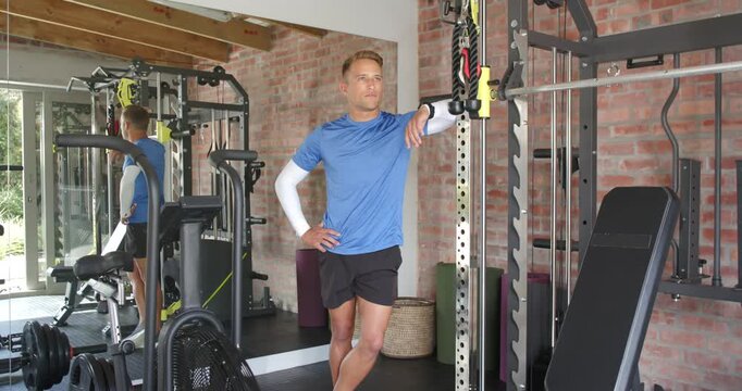 Man leaning on squat rack at gym, shifting weight, glancing mirror, checking wrist device for rest
