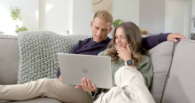 Couple sitting on grey sofa, man with silver laptop explaining screen content while woman reacting