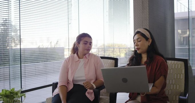 Diverse female coworkers sitting in lounge, right colleague holding laptop, explaining project
