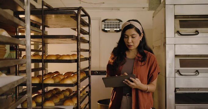Midadult woman in rust shirt scanning racks using tablet logging fresh rolls for quality at bakery