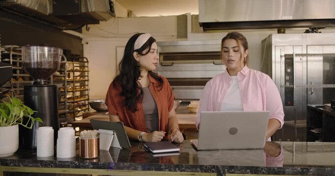 Diverse women left gesturing, right typing laptop or tablet at bakery counter checking orders