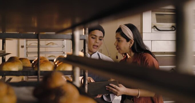 Diverse female chef and manager inspecting fresh loaves on racks in bakery, comparing tablet