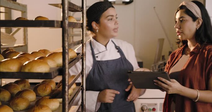 Female bakery team at racks, baker in chef coat pointing while manager using tablet checking output