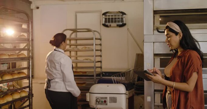 Diverse female crew manager in bakery using tablet, chef checking bread on tall rack, counting