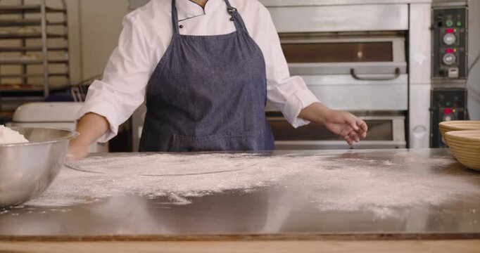 Female chef in jacket apron reaching mix bowl, dusting, smoothing flour on prep table for dough