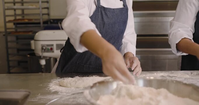 Diverse female bakers in jackets sprinkling flour from bowl, folding dough at bakery for baking