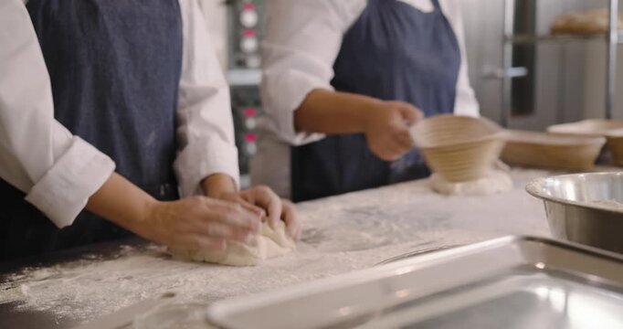 Diverse female coworkers after mixing, in chef coats shaping dough and dusting baskets for proofing