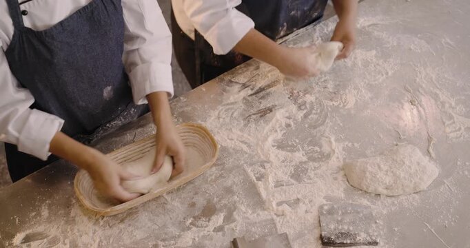 Female bakers shaping dough for proof on steel bench wearing chef coats, placing loaves in banneton
