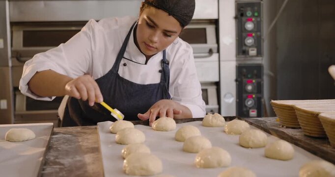 Diverse female bakers in chef whites brushing gloss with yellow brush, preparing buns for baking